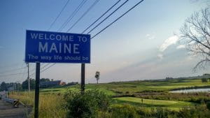 Clear skies with a few scattered clouds on the right side of the image. Blue street sign with white letters that say “Welcome to Maine. The way life should be”. A landscape with fields, lawns and a small part that can be seen as a water surface. There are cables that stretch from one electric post to another.