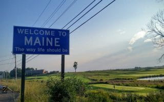 Clear skies with a few scattered clouds on the right side of the image. Blue street sign with white letters that say “Welcome to Maine. The way life should be”. A landscape with fields, lawns and a small part that can be seen as a water surface. There are cables that stretch from one electric post to another.