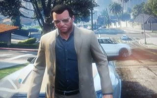 Grand Theft Auto character Michael De Santa in a beige suit with button down shirt and sunglasses standing in front of his default vehicle