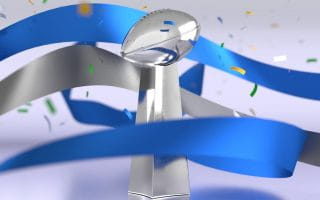 The silver Lombardi Trophy awarded to the Super Bowl Champion team surrounded by multicolored confetti and blue and gray ribbons