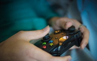 Someone holding a black Xbox One wireless video game controller with its trademark yellow Y, blue X, green A, and red B buttons.