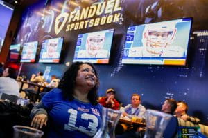 Woman in a New York Giants jersey at a New Jersey sportsbook during the Super Bowl with Tom Brady on the TV screens.