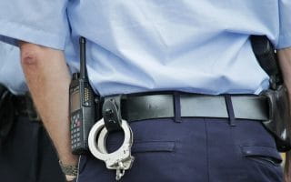 The rear view of a police officer wearing navy blue pants and a sky blue shirt in addition to handcuffs and a communication device on his belt
