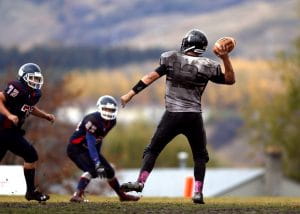 A quarterback in a dirty white uniform with black pants attempting to throw a pass while being defended by two players on the opposing team in navy blue uniforms