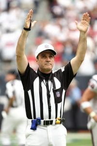 An NFL referee wearing the customary black and white striped shirt along with a white hat and white pants and his yellow penalty flag and blue fumble bag on his belt