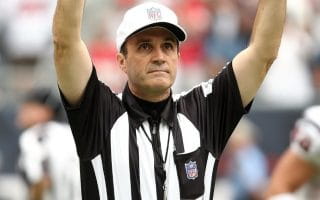 An NFL referee wearing the customary black and white striped shirt along with a white hat and white pants and his yellow penalty flag and blue fumble bag on his belt