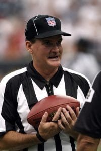 An NFL referee wearing the customary black and white striped shirt along with a black hat and whistle wrapped around his wrist while cradling an NFL football.  