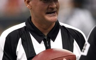 An NFL referee wearing the customary black and white striped shirt along with a black hat and whistle wrapped around his wrist while cradling an NFL football.
