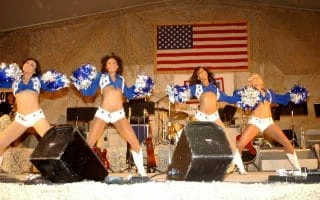 Dallas Cowboy cheerleaders in white shorts and white and blue blouses with white and blue pompoms dancing with an American flag in the background.