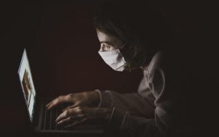 A woman with surgery mask typing on a laptop