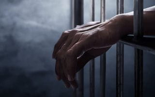 Person’s Hands Behind Bars Over a Dark Background 