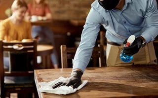 A Waiter with Mask and Gloves Cleaning a Table in a Caffee Using Detergent